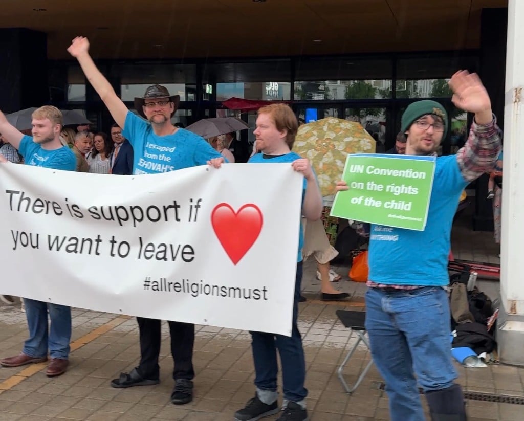 Peaceful ExJW protesters holding a banner reading 'There is support if you want to leave' and a sign about the UN Convention on the Rights of the Child, #allreligionsmust